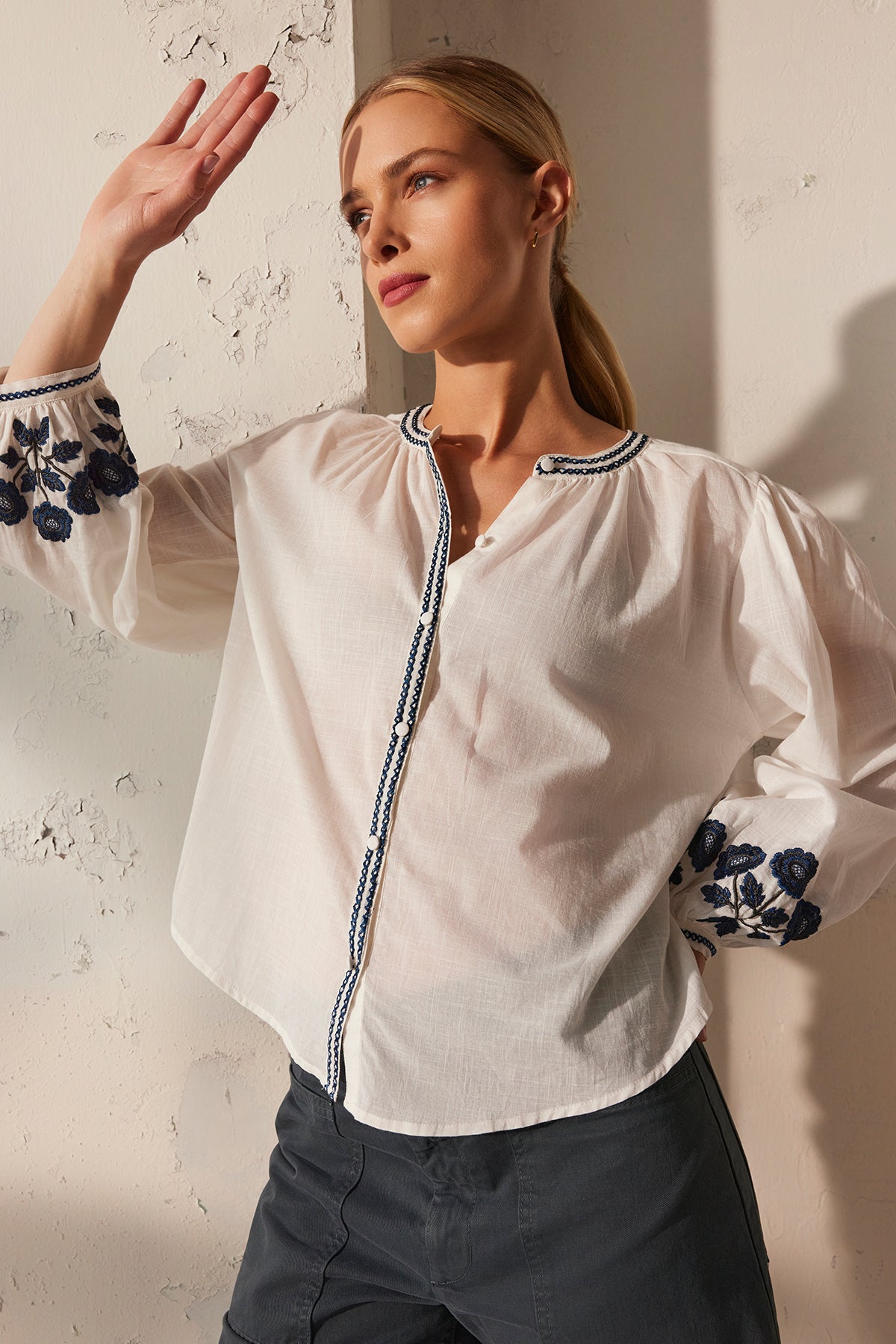 Wearing the RAHWA TOP by Velvet by Graham & Spencer, a person poses against a textured wall, arm raised. The lightweight cotton top features floral embroidery on the sleeves and a button-front design.-38866415780033