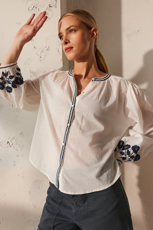 Wearing the RAHWA TOP by Velvet by Graham & Spencer, a person poses against a textured wall, arm raised. The lightweight cotton top features floral embroidery on the sleeves and a button-front design.