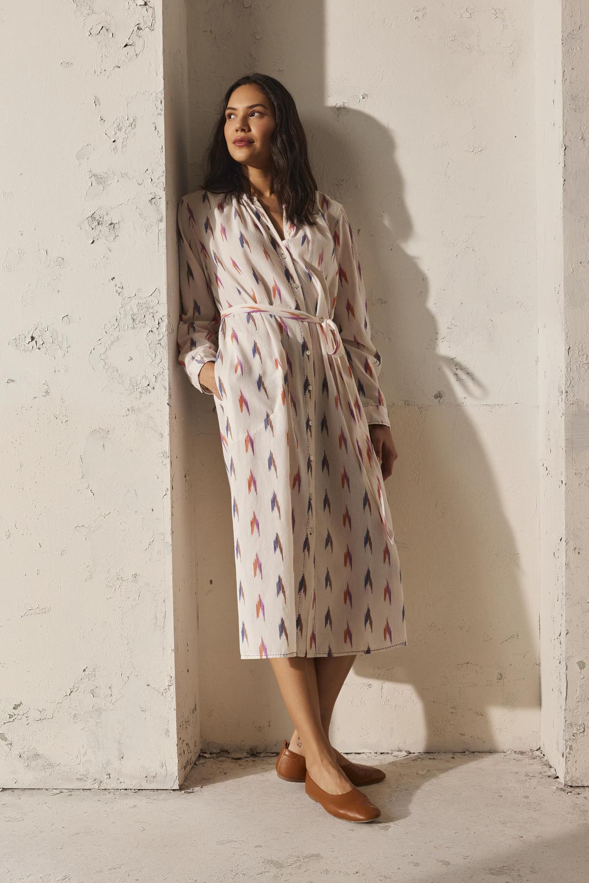 Wearing the ALTA DRESS by Velvet by Graham & Spencer, a woman in a lightweight cotton ikat weave dress leans against a textured wall as sunlight casts shadows. Her hands rest in her pockets, complementing her brown shoes.-38512666181825