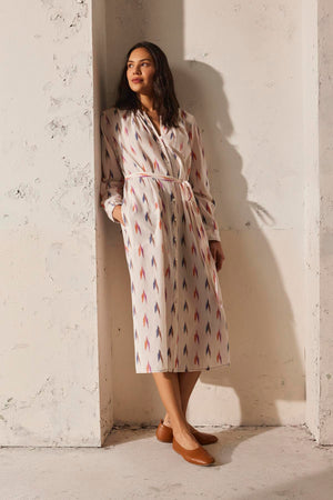 Wearing the ALTA DRESS by Velvet by Graham & Spencer, a woman in a lightweight cotton ikat weave dress leans against a textured wall as sunlight casts shadows. Her hands rest in her pockets, complementing her brown shoes.