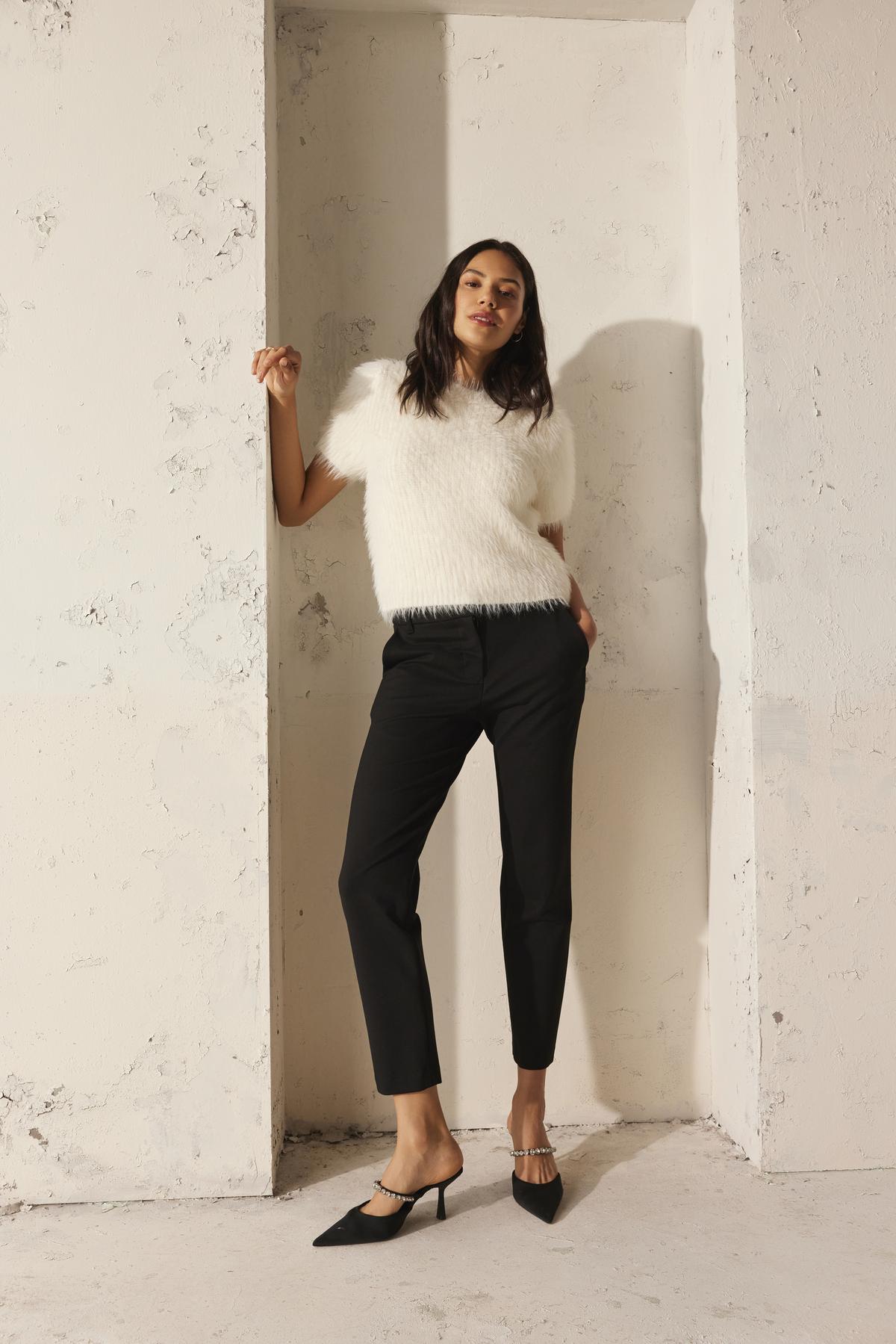 A woman wearing the EILA FEATHER YARN SWEATER from Velvet by Graham & Spencer and black pants leans against a textured, light-colored wall while donning black heels.-38246411763905