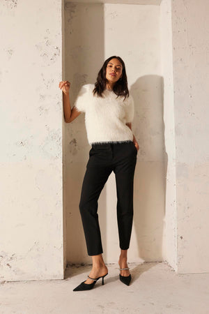 A woman wearing the EILA FEATHER YARN SWEATER from Velvet by Graham & Spencer and black pants leans against a textured, light-colored wall while donning black heels.