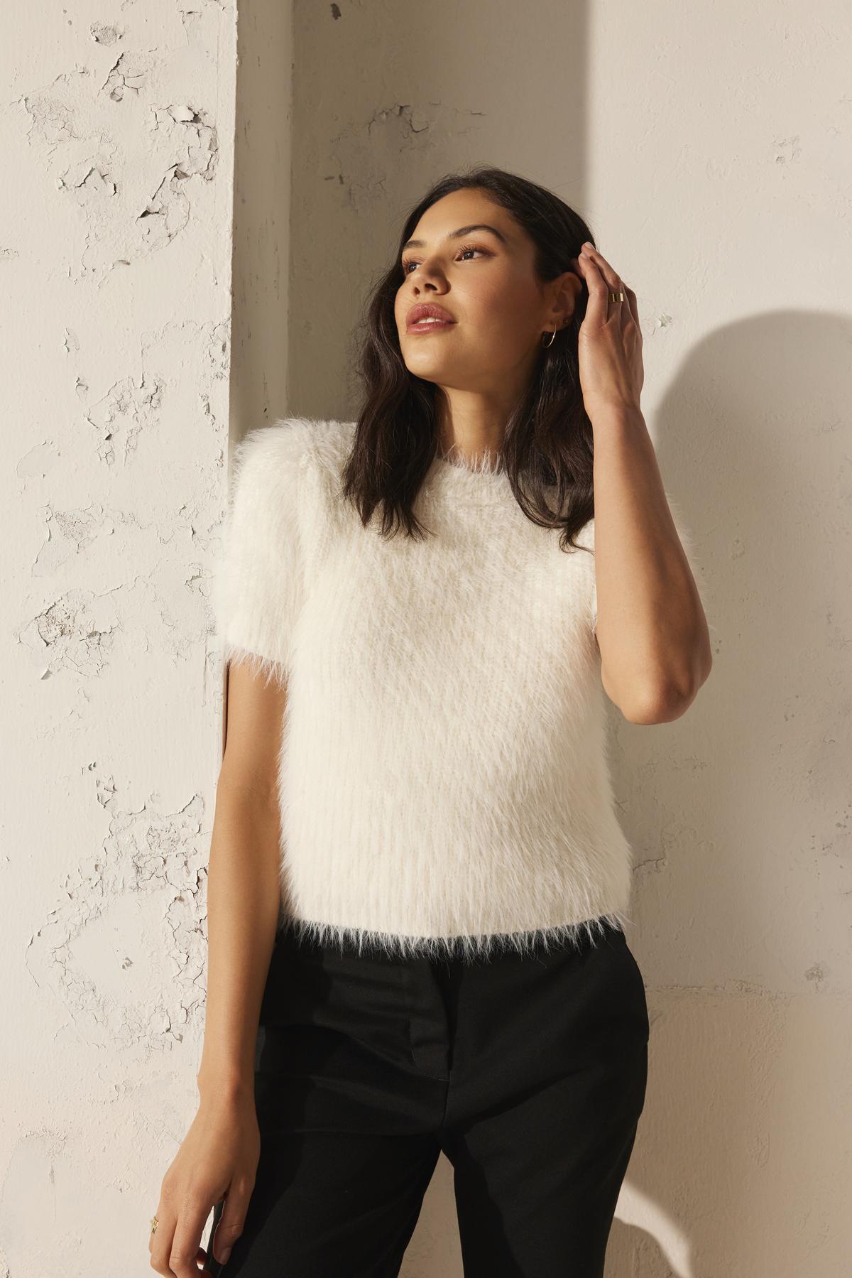 A woman stands against a textured wall, wearing the EILA FEATHER YARN SWEATER by Velvet by Graham & Spencer, paired with black pants, with her hand touching her hair.-38246411796673