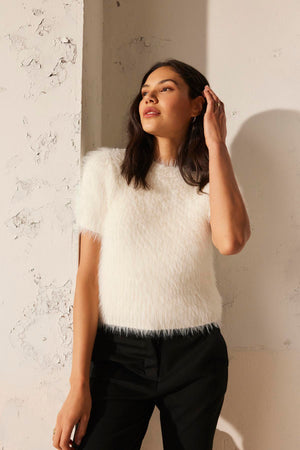 A woman stands against a textured wall, wearing the EILA FEATHER YARN SWEATER by Velvet by Graham & Spencer, paired with black pants, with her hand touching her hair.
