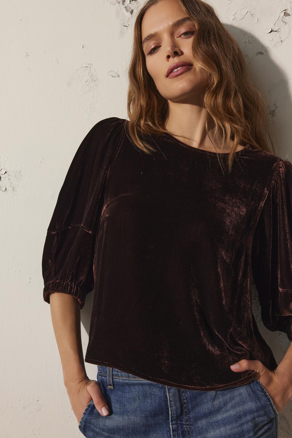 A woman with wavy hair poses against a textured wall, wearing the luxurious NANCY SILK VELVET TOP by Velvet by Graham & Spencer, paired with denim jeans.