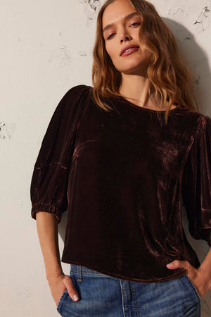 A woman with wavy hair poses against a textured wall, wearing the luxurious NANCY SILK VELVET TOP by Velvet by Graham & Spencer, paired with denim jeans.