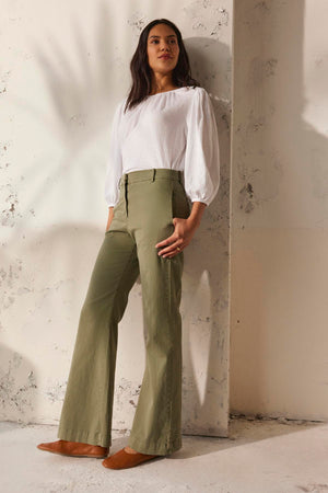 A person wearing a white blouse and the olive green COBIE PANT by Velvet by Graham & Spencer leans against a textured wall in soft lighting.