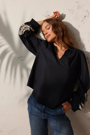 A woman in a MARTINE TOP by Velvet by Graham & Spencer and jeans stands against a textured wall with shadow patterns, eyes closed, one arm raised.