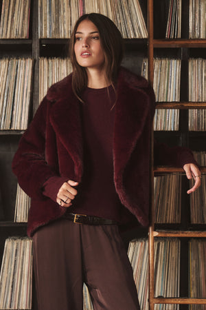 A woman wears the CAROLYN FAUX FUR JACKET by Velvet by Graham & Spencer—burgundy with a lapel collar—paired with brown pants, standing before shelves lined with vinyl records.