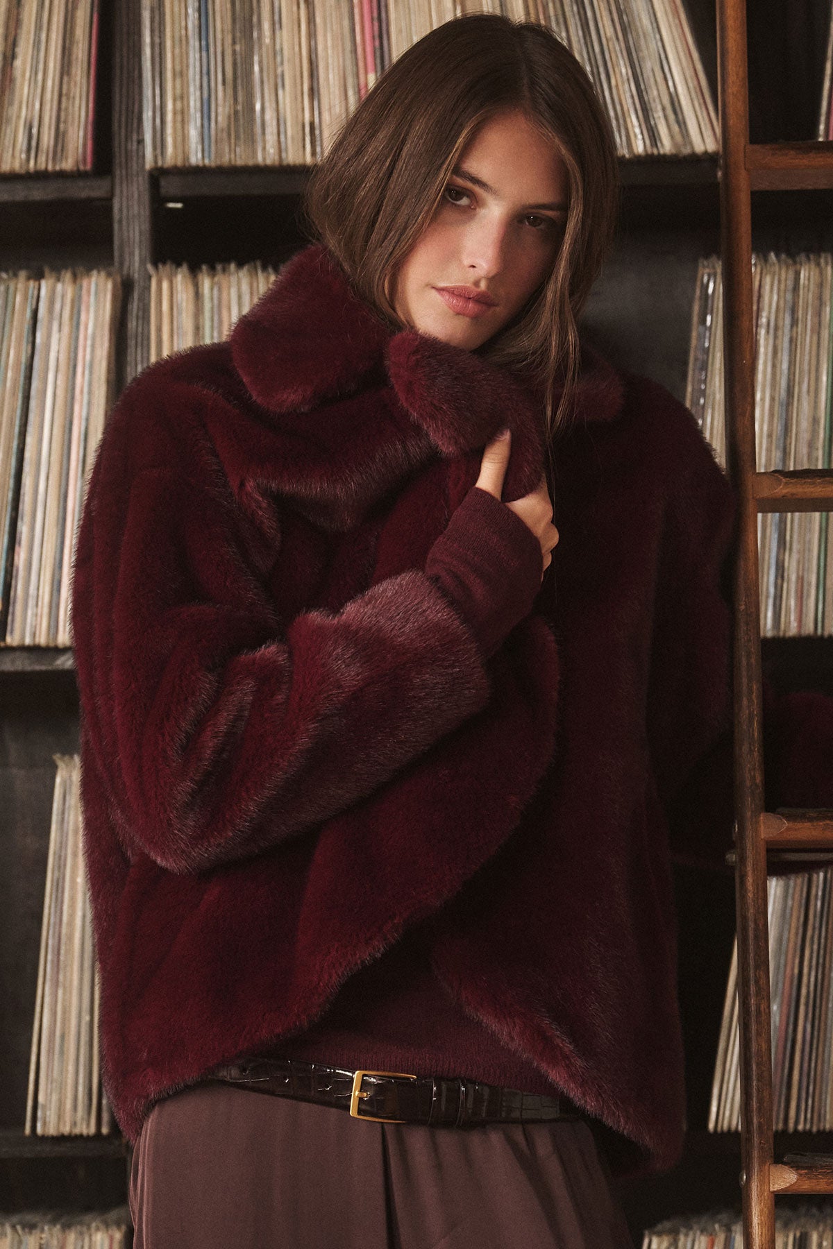 A person wearing the CAROLYN FAUX FUR JACKET by Velvet by Graham & Spencer with a chic lapel collar stands before shelves of vinyl records, holding the collar and looking at the camera.