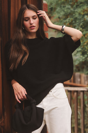 A woman stands outdoors against a wooden wall, wearing a loose black top and white pants, holding the ROBYN SATIN BAG by Velvet by Jenny Graham, with greenery in the background.