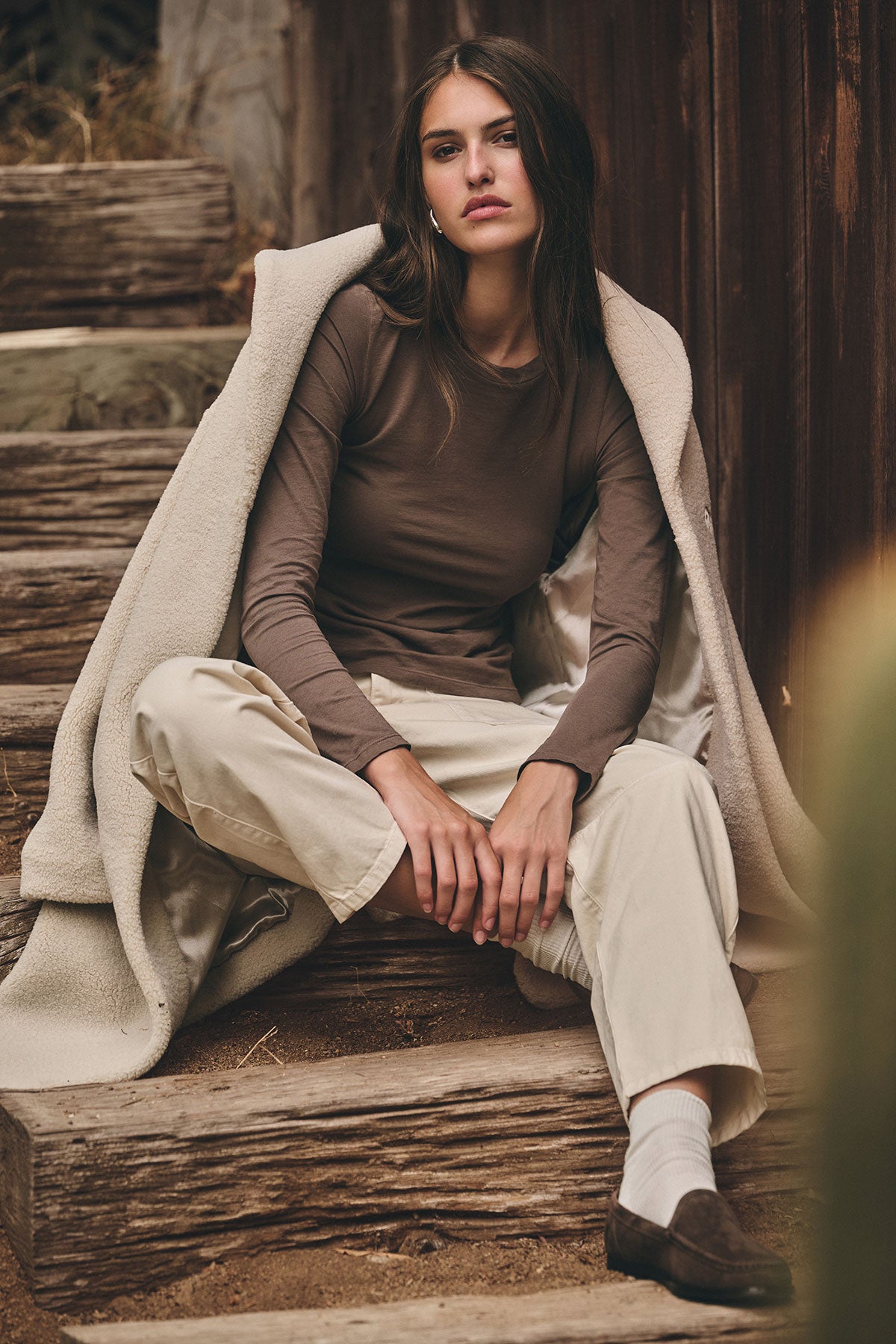 A woman sits on wooden steps, wearing the Velvet by Graham & Spencer ZOFINA TEE—an ultra-soft, fitted long sleeve top with a classic crew neck—paired with light beige pants, a draped light coat, white socks, and dark shoes.