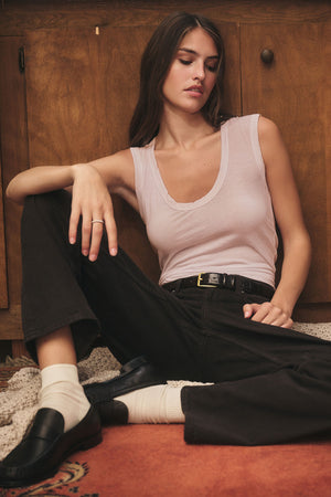 A woman sits on the floor in the ESTINA TANK TOP by Velvet by Graham & Spencer, paired with black pants, white socks, and black loafers as she leans against a wooden cabinet.