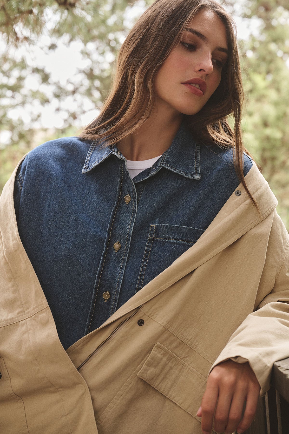 Wearing the ROSA JACKET by Velvet by Graham & Spencer over a blue denim shirt, a woman stands outdoors among trees, capturing an effortless everyday style.