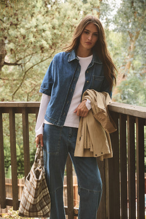 A woman stands on a wooden deck amid trees, wearing Velvet by Graham & Spencer's FARAH DENIM PANT—cropped with patch pockets and a curved leg silhouette—paired with a short-sleeved denim shirt, white top, plaid bag, and beige coat.