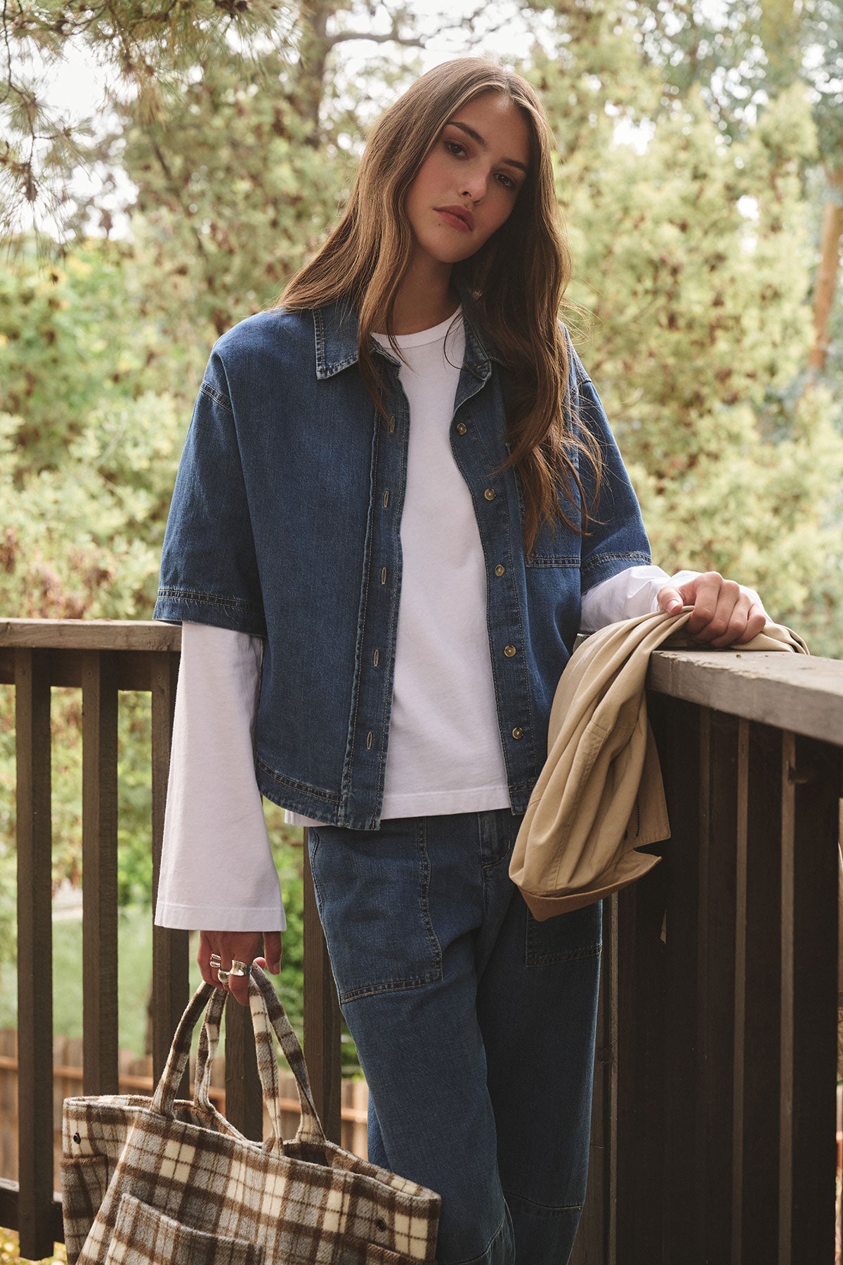 A woman stands outdoors on a wooden deck, wearing the VEE TEE by Velvet by Graham & Spencer under a denim jacket with jeans, holding a plaid bag and beige coat, with trees in the background.
