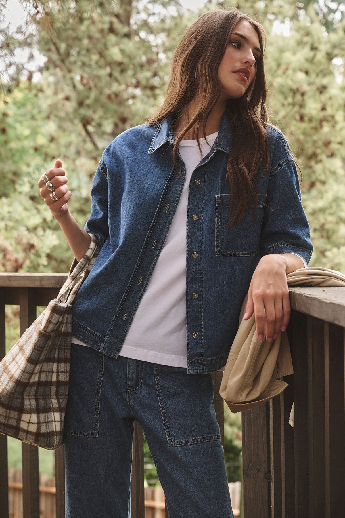 Wearing the KADDY DENIM TOP by Velvet by Graham & Spencer, a woman stands on a wooden balcony with jeans, holding a plaid bag and beige jacket, framed by trees in the background.-39839618760897