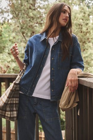 Wearing the KADDY DENIM TOP by Velvet by Graham & Spencer, a woman stands on a wooden balcony with jeans, holding a plaid bag and beige jacket, framed by trees in the background.