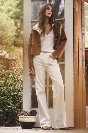 A woman stands before a glass door wearing Velvet by Graham & Spencer’s WINONA POINTELLE HENLEY under a brown jacket with high-waisted white pants, one hand in her pocket and an apple in the other.