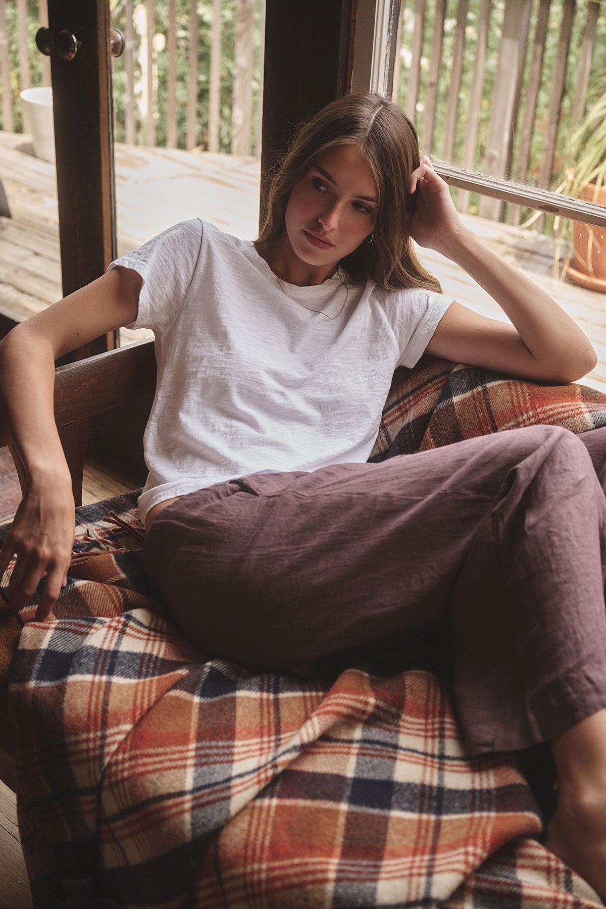 A woman in a white tee and Velvet by Graham & Spencer's LOLA LINEN PANT sits on a plaid blanket-covered couch, resting her head on one hand near a window.-39839701663937