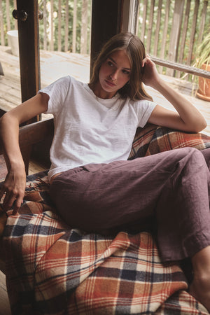 A woman in a white tee and Velvet by Graham & Spencer's LOLA LINEN PANT sits on a plaid blanket-covered couch, resting her head on one hand near a window.