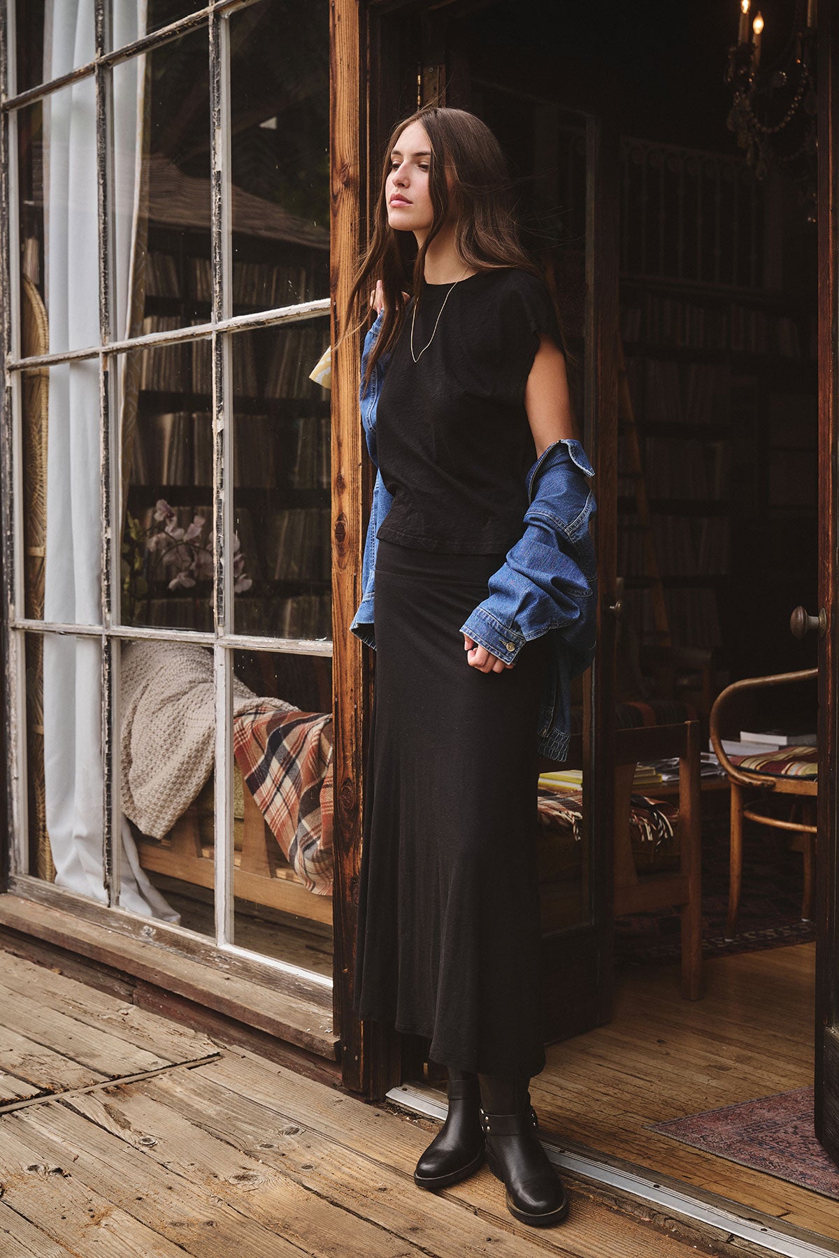 A woman stands in a doorway wearing the STERLING TEE by Velvet by Graham & Spencer, styled with black boots, a blue denim jacket draped over her arms, and a necklace; wooden floors and bookshelves are in the background.-39840098320577