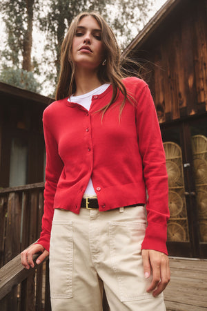A woman stands outdoors in the VIOLETTE CARDIGAN by Velvet by Graham & Spencer—a cropped, red cotton-cashmere layer over a white top and beige pants, ideal for lightweight layering against a backdrop of wooden buildings.