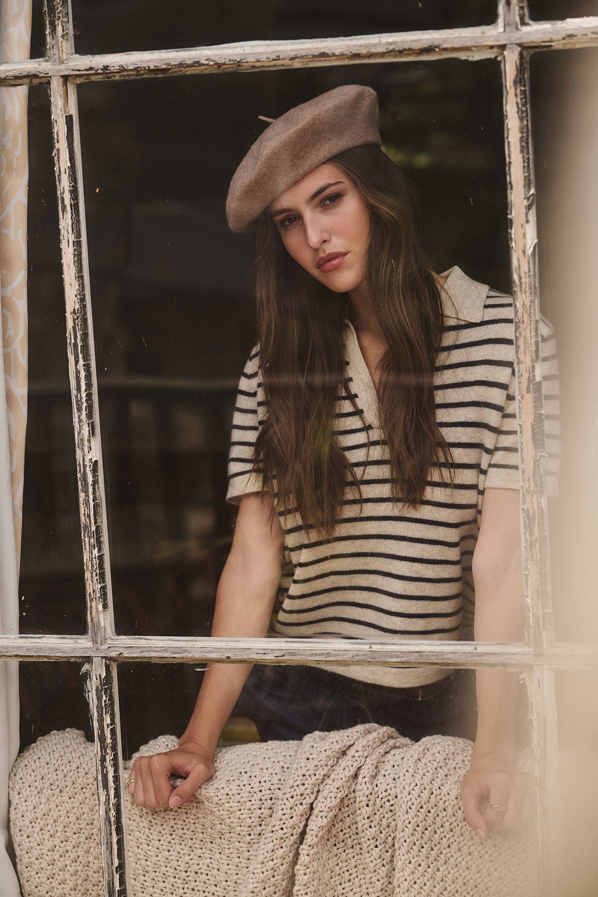 A woman in a Velvet by Graham & Spencer CLASSIC BERET and striped shirt stands behind a window, her hand resting on a knitted blanket draped over the couch.