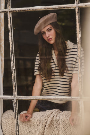 A woman in a Velvet by Graham & Spencer CLASSIC BERET and striped shirt stands behind a window, her hand resting on a knitted blanket draped over the couch.