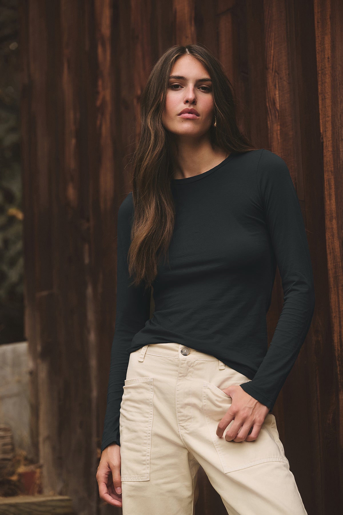 A woman with long brown hair, wearing the ZOFINA TEE by Velvet by Graham & Spencer and beige pants, stands in front of a wooden wall.