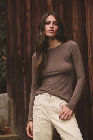 A woman with long brown hair wears the ZOFINA TEE by Velvet by Graham & Spencer—an ultra-soft, fitted long sleeve crewneck—paired with beige pants and stands in front of a wooden wall.