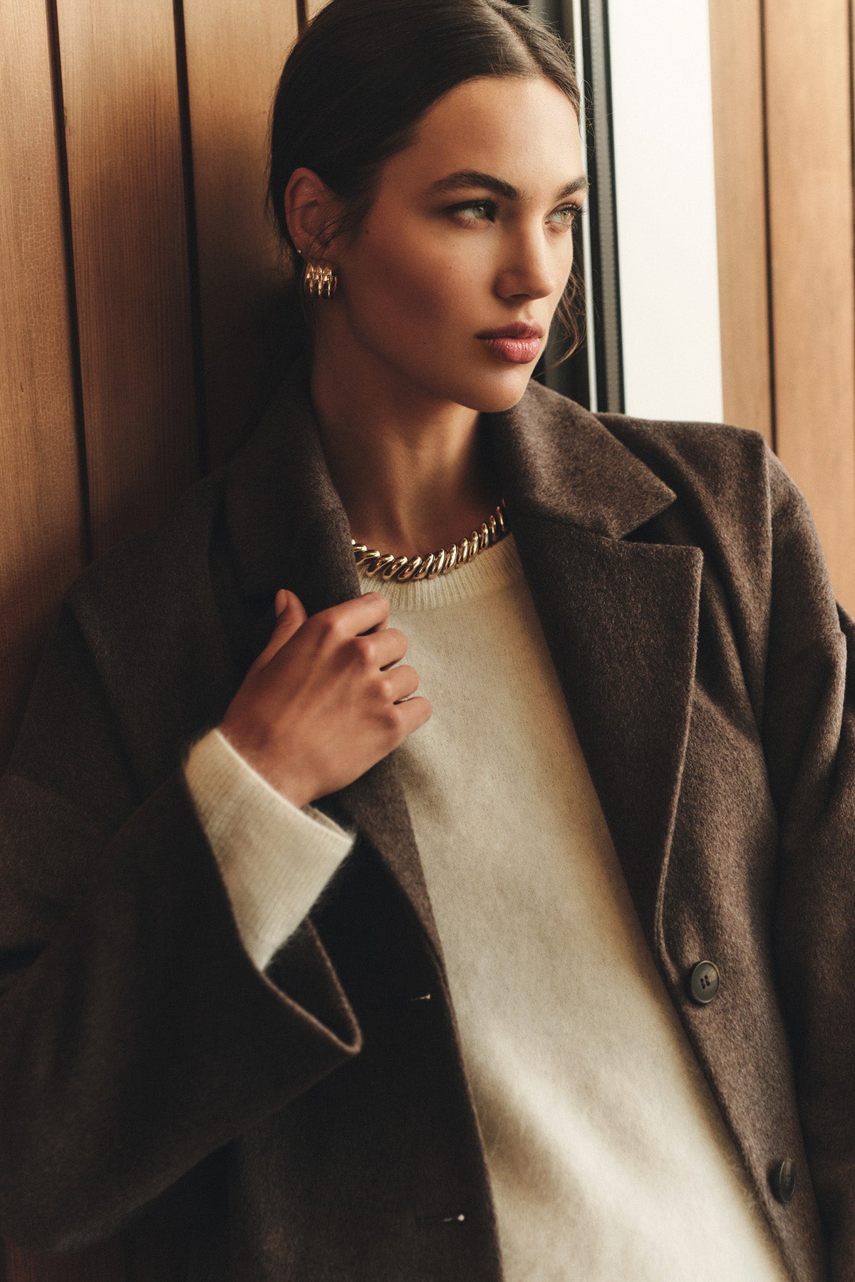 Wearing the LAFAYETTE COAT by Velvet by Graham & Spencer over a white sweater, a person stands by a wooden wall, looking away from the camera and accessorized with gold jewelry.-40263652049089