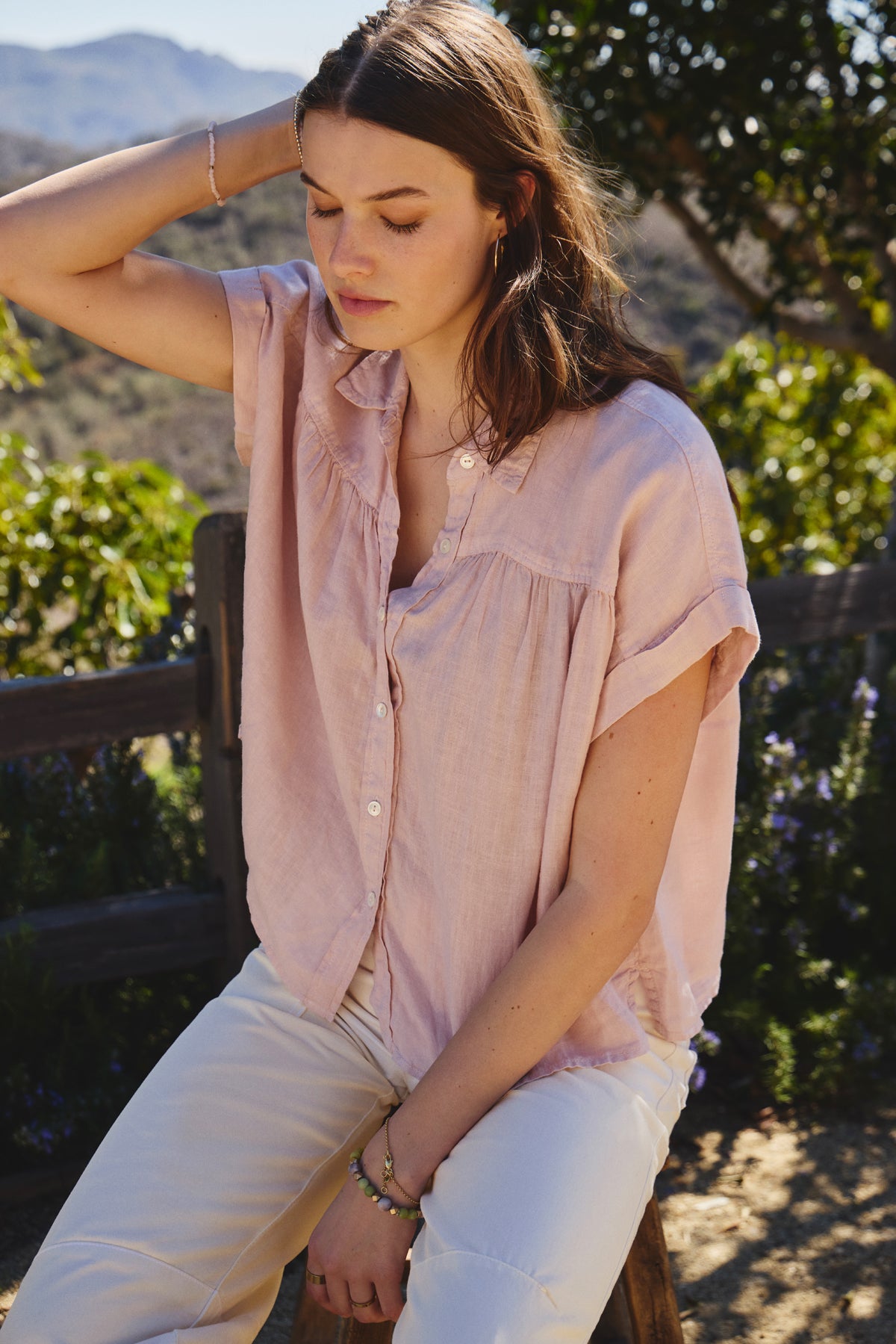 A person sits outdoors on a wooden bench wearing the ARIA LINEN TOP from Velvet by Graham & Spencer, which features a light pink hue and button-front design. The classic collar enhances its elegance, while greenery and distant mountains bask under clear skies in the background.