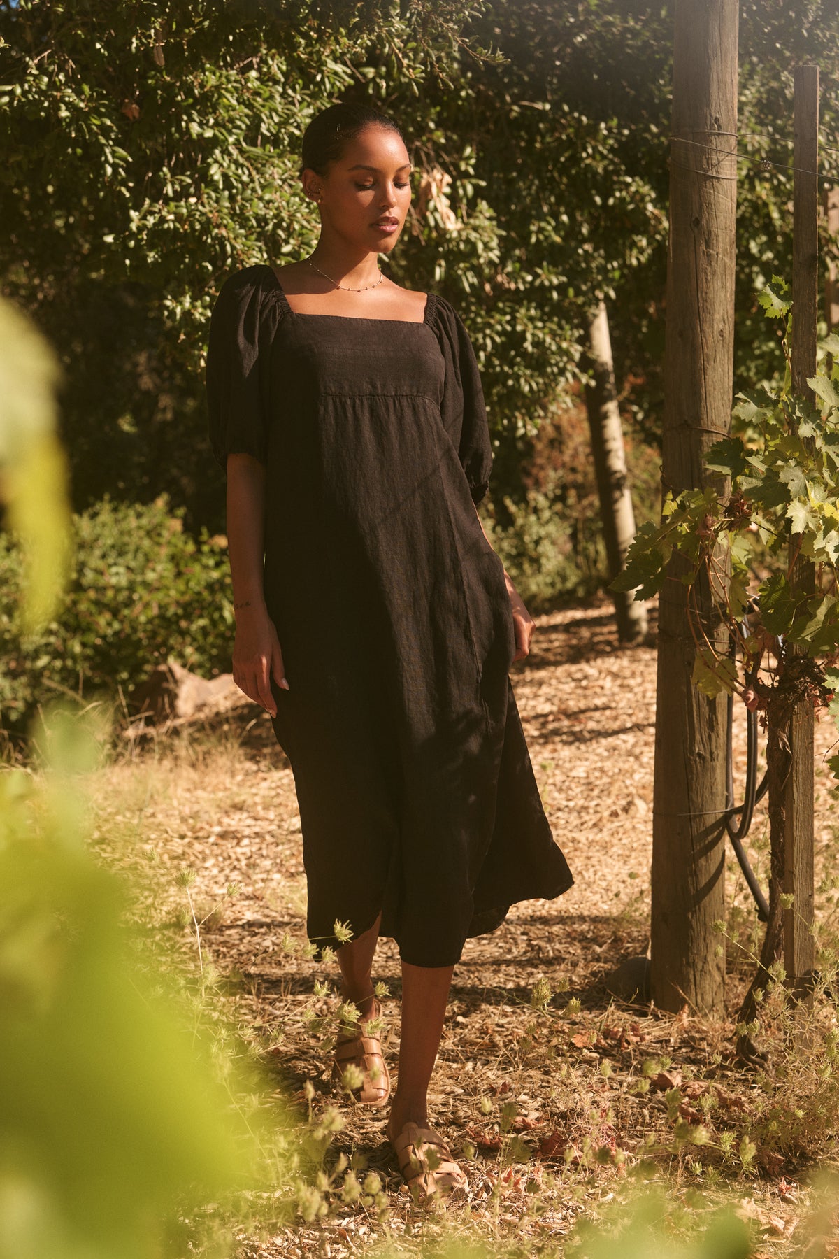 A person wearing the BROOKE LINEN DRESS by Velvet by Graham & Spencer strolls along a sunlit path with greenery in the background.-38643528040641