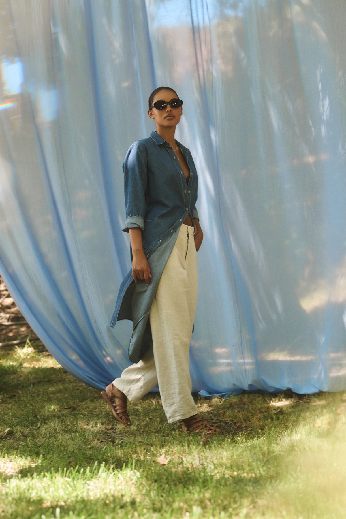 A person in an IZMIR CHAMBRAY DRESS by Velvet by Graham & Spencer, paired with white pants and sunglasses, walks on grass against a large blue fabric backdrop.-38820646322369
