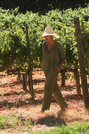 A person dressed in Velvet by Graham & Spencer's GINI LINEN PANT and a wide-brimmed hat strolls through a vineyard under the sunny sky.