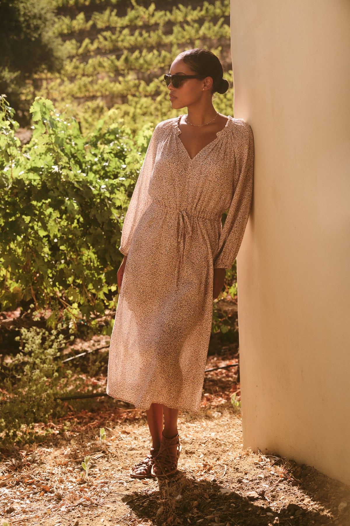 A woman in a LIMA Printed Crinkle Cotton Gauze Dress by Velvet by Graham & Spencer, wearing sunglasses and leaning against an outdoor wall, surrounded by greenery.-38820676272321