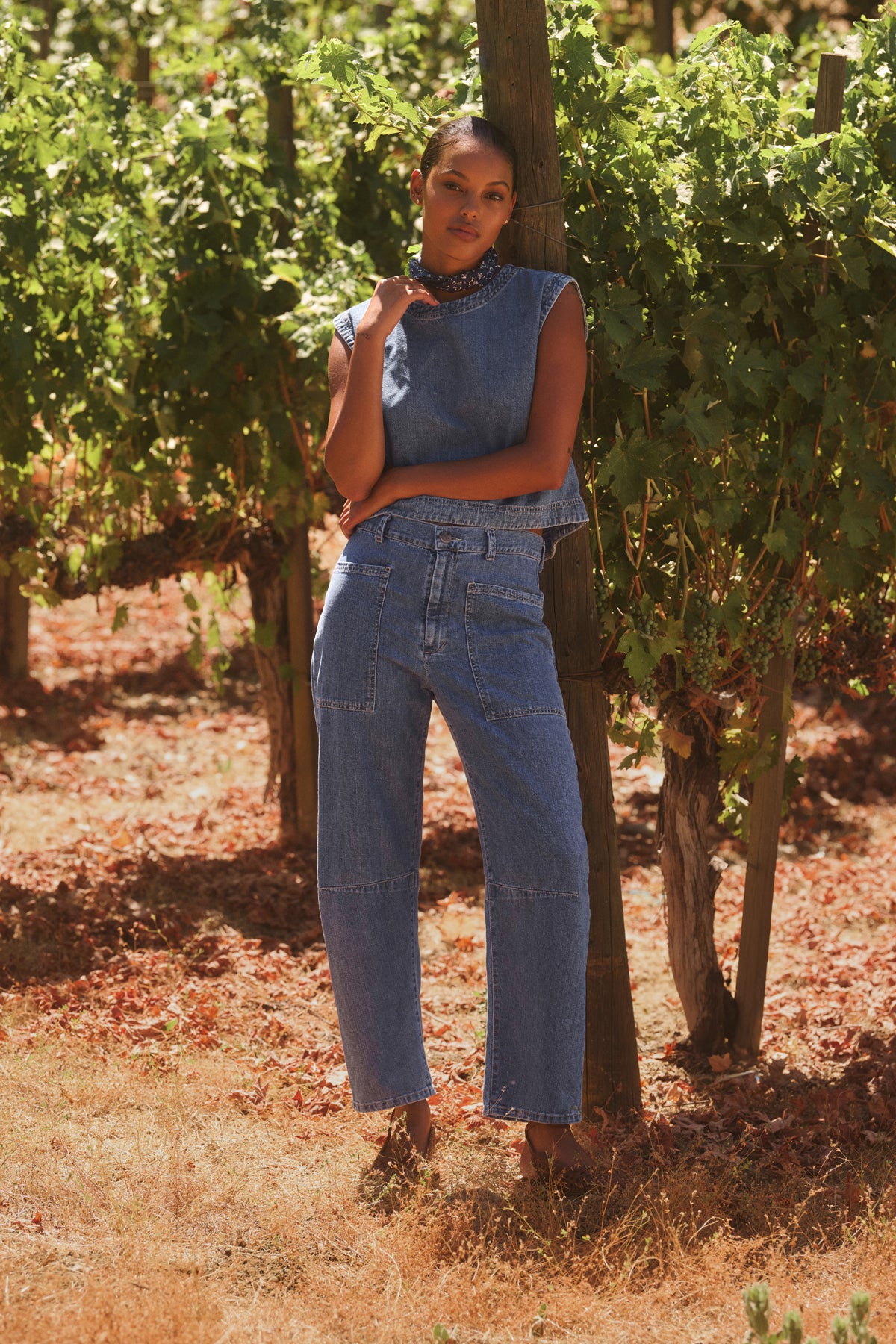 A person in Velvet by Graham & Spencer's FARAH DENIM PANTS stands against a wooden post in a vineyard, surrounded by green grapevines.-38820625776833