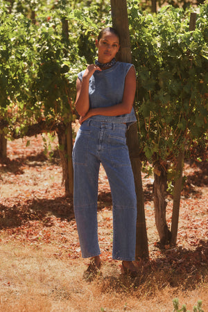 A person in Velvet by Graham & Spencer's FARAH DENIM PANTS stands against a wooden post in a vineyard, surrounded by green grapevines.