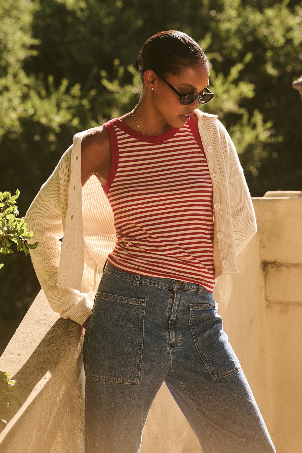 A person wearing the LUA TANK TOP by Velvet by Graham & Spencer, styled with sunglasses and a white cardigan, leans on a concrete railing with sunlight and greenery in the background.-38820722245825