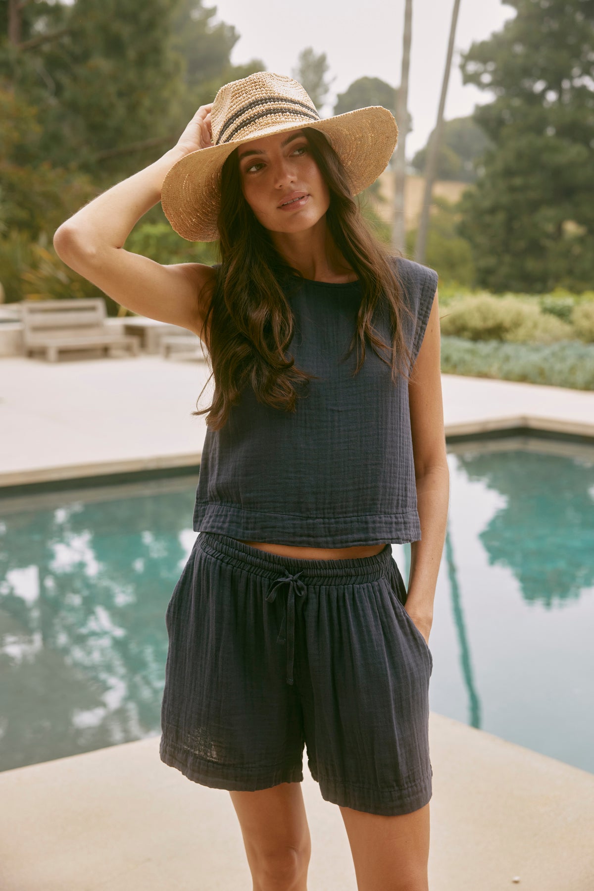 A woman wearing casual dark TIBBY COTTON GAUZE SHORT by Velvet by Graham & Spencer with a drawstring elasticized waist and a wide-brimmed straw hat poses with one hand on her hat. She stands by a pool with trees and outdoor furniture in the background.