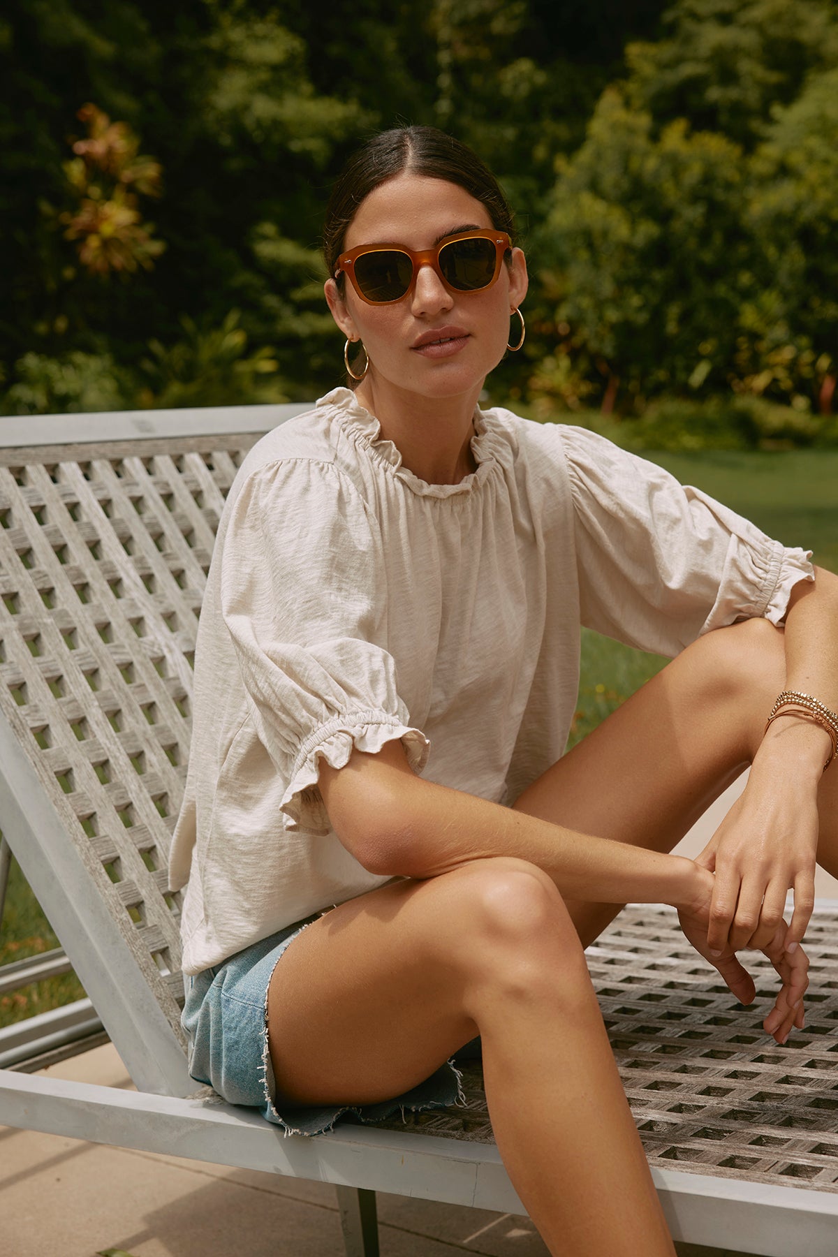 A woman wearing sunglasses,  the CARLENE TEE by Velvet by Graham & Spencer, and denim shorts sits on a metal outdoor chair in a garden, enjoying the relaxed fit of her breathable cotton outfit.