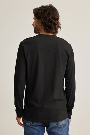 A man with wavy hair, wearing a black ALVARO HENLEY by Velvet by Graham & Spencer and blue jeans, looks to the side against a plain background.