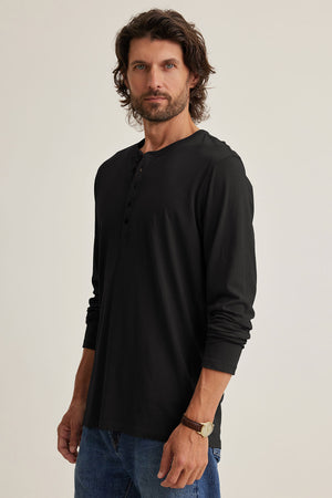 A man with shoulder-length hair and a beard showcases his casual style in an ALVARO HENLEY from Velvet by Graham & Spencer, made from soft Pima jersey, against a plain background.