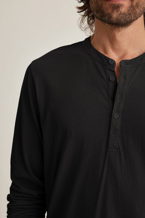 The photo shows someone wearing a Velvet by Graham & Spencer ALVARO HENLEY, featuring black Pima jersey fabric, long sleeves, and four buttons. It's captured from the shoulders to the upper chest for a stylish layering look.