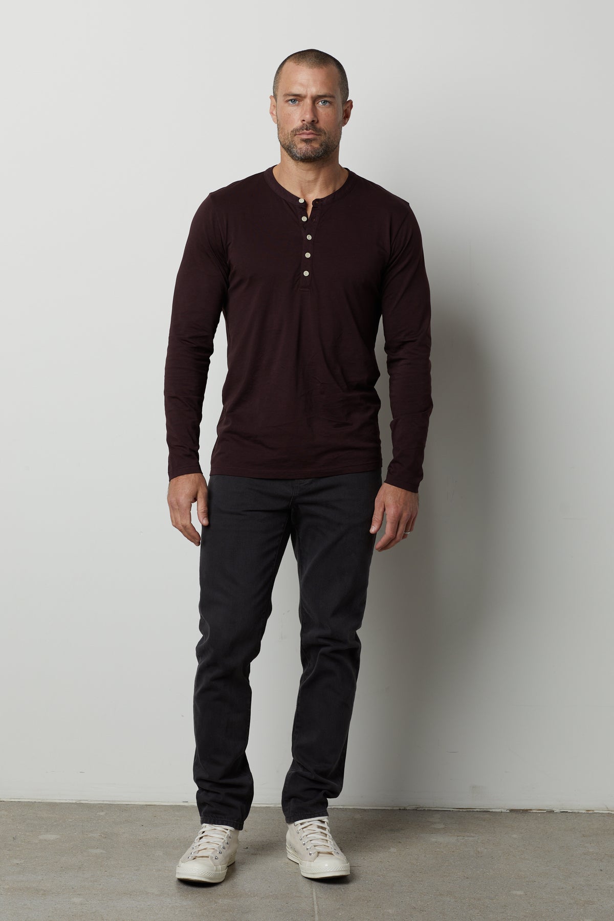 A man stands against a plain white background wearing a dark, lightweight ALVARO HENLEY by Velvet by Graham & Spencer, dark pants, and white sneakers.
