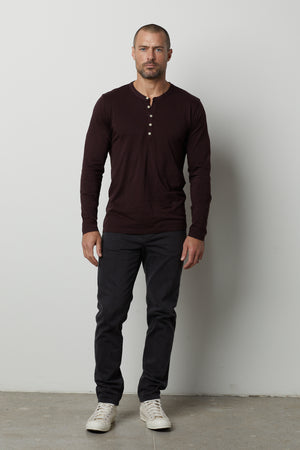A man stands against a plain white background wearing a dark, lightweight ALVARO HENLEY by Velvet by Graham & Spencer, dark pants, and white sneakers.