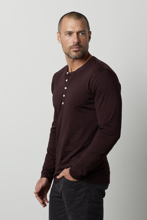 A man with a shaved head and short beard is wearing a long-sleeved, dark red ALVARO HENLEY by Velvet by Graham & Spencer and black jeans, standing against a plain, light gray background.