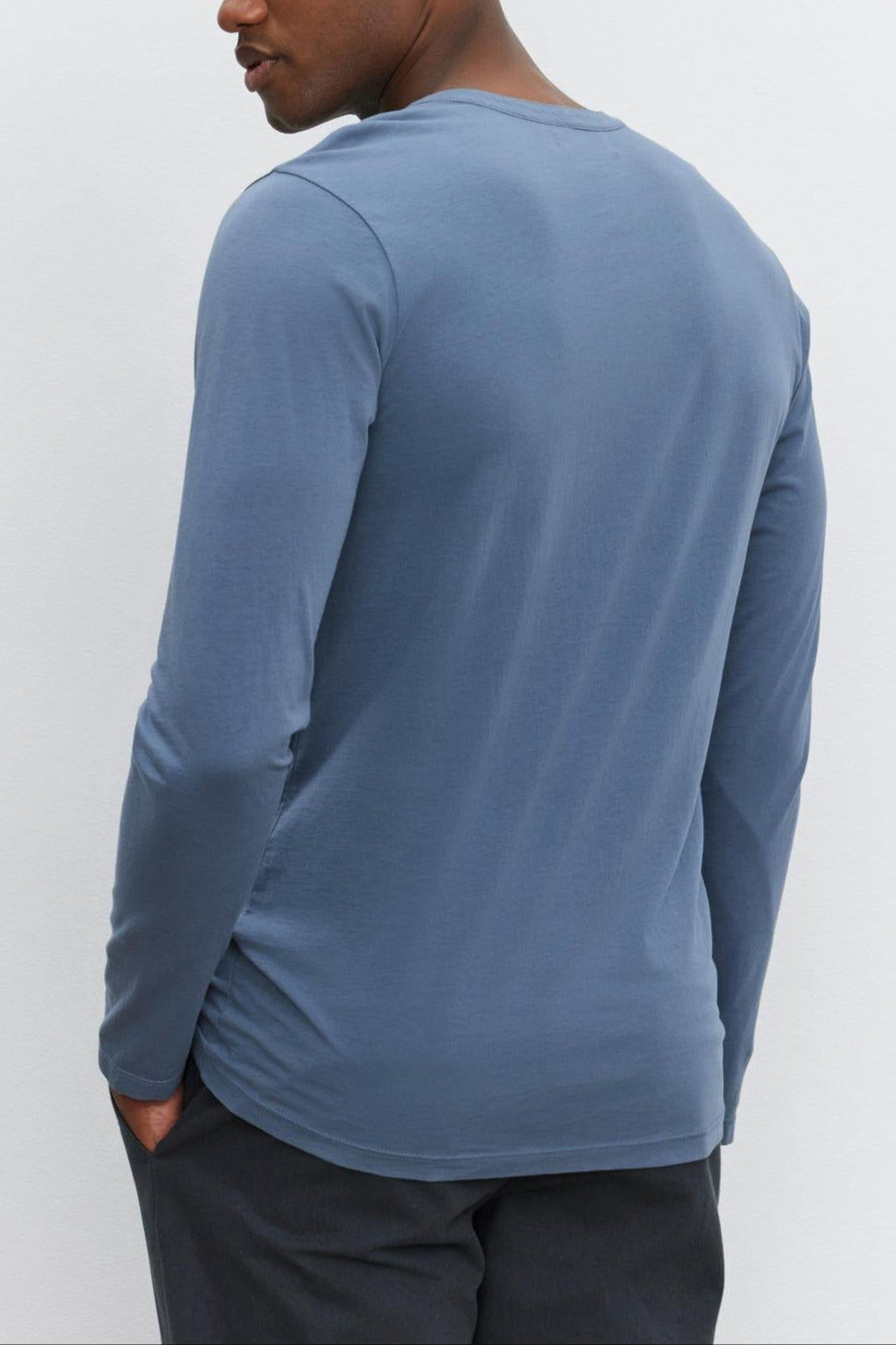 A person wearing the ALVARO HENLEY, a long-sleeved blue lightweight shirt by Velvet by Graham & Spencer, paired with dark pants, is standing with their back to the camera, looking to the side.
