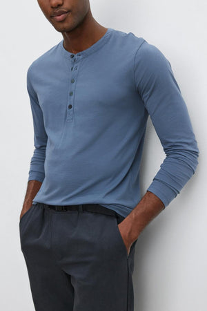 A man with short hair stands casually with hands in pockets, wearing the ALVARO HENLEY by Velvet by Graham & Spencer— a lightweight blue henley shirt that's perfect for layering. He pairs it with dark pants, all set against a plain white background.